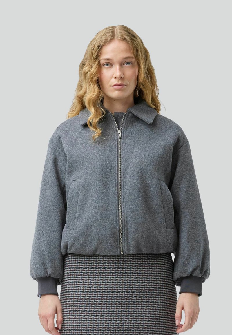 Una donna con lunghi capelli mossi indossa una giacca bomber grigia su una gonna a scacchi, posando su uno sfondo grigio uniforme.