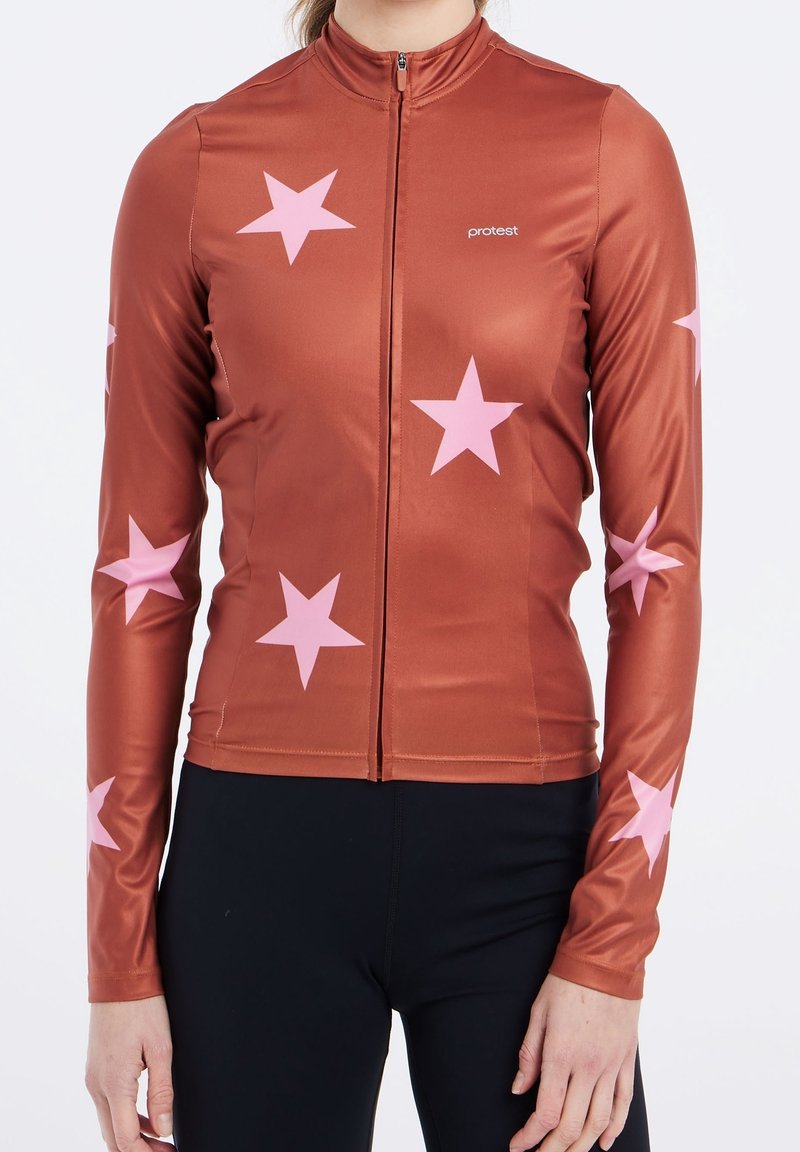 Maillot de cyclisme à manches longues couleur rouille avec des étoiles roses, fermeture éclair intégrale à l'avant, logo "protest" sur la poitrine, porté avec un pantalon noir.