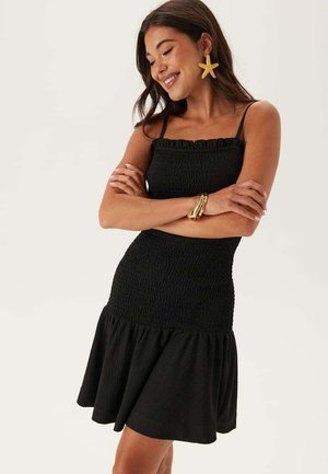Sort kjole med smockeffekt på toppen, flæseudskæring og udvidet skørt. Lavet af et struktureret stof med justerbare spaghettistropper.