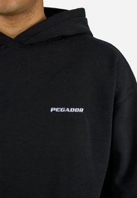 Schwarzer Kapuzenpullover mit weißem "PEGADOR"-Logo, das auf der linken Brust gestickt ist, getragen von einer Person, die vom Hals bis zur Schulter zu sehen ist.