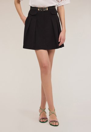 Falda negra plisada con bolsillos y un cinturón negro con hebilla dorada. La modelo lleva sandalias con tiras en verde y rosa. Fondo claro.