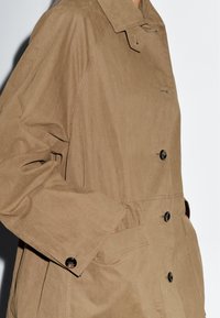 Beige trenchcoat van lichtgewicht stof, met een puntige kraag, grote knopen en zijzakken, met een strakke, op maat gemaakte uitstraling.