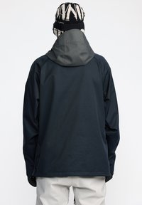 Veste coupe-vent en matériau bleu foncé avec une capuche grise. Elle possède des manches longues et une coupe décontractée, avec une texture lisse et un minimalisme dans les détails.
