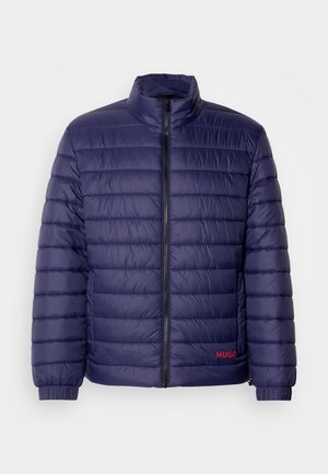 Mörkblå vadderad jacka med horisontellt quiltat mönster, hög krage, dragkedja framtill och röd logodetalj på vänster sida.
