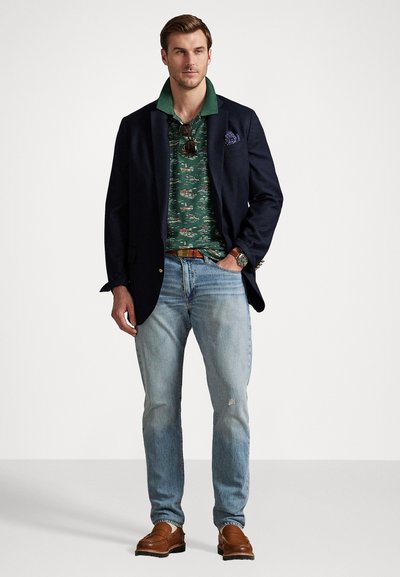 Jūras flotes apģērbs ar zaļganrakstu polo, gaiši zili džinsi, brūni loafersi, rokas pulkstenis, saulesbrilles uz apkakles un kabatas kvadrāts žaketē.