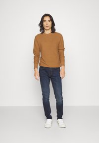 Maglione marrone a coste con collo rotondo, abbinato a jeans slim-fit blu scuro e sneakers bianche, esposto su uno sfondo neutro.