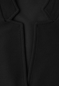 Gros plan d’un tissu noir texturé avec une couture centrale et un motif découpé angulaire, montrant des motifs de tricot détaillés.
