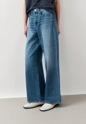 Henkilö pukeutuneena leveälahkeisiin sinisiin farkkuihin, mustaan toppiin ja valkoisiin neliökärkisiin kenkiin, seisoo yksivärisen vaalean taustan edessä.