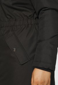 Close-up van een zwarte winterjas met elastische taille, klepzak met drukknoop en een afgewerkte manchetrand bij een hand.