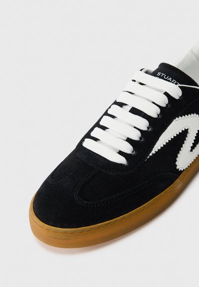 Baskets en suede noir avec des lacets blancs, un design en zigzag blanc sur le côté et une semelle en caoutchouc marron sur un fond clair.