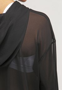 Chaqueta ligera y transparente con capucha de color negro y corte relajado. Presenta una textura suave y un brillo sutil que resalta el diseño en capas.