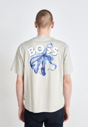 Mężczyzna w jasnoszarej koszulce z niebieską grafiką ośmiornicy i dużym białym napisem "BOSS" na plecach, stojący tyłem do kamery.