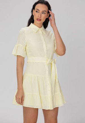 Robe jaune clair avec un col, des manches courtes à volants, un devant boutonné et une taille à nouer. Jupe évasée avec des rayures verticales.