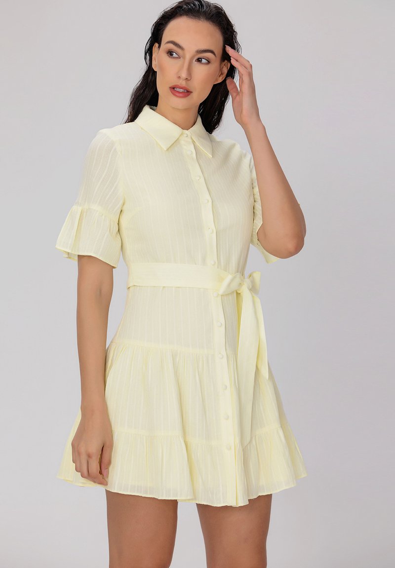 Robe jaune clair avec un col, des manches courtes à volants, un devant boutonné et une taille à nouer. Jupe évasée avec des rayures verticales.