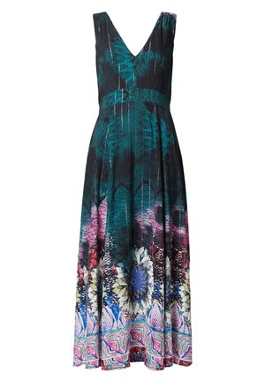 Frau in einem ärmellosen Maxikleid mit blauen und lila Blumenmustern, weißen Sneakern, die eine weiße Jacke und eine cremefarbene Umhängetasche mit dem Schriftzug "Love" hält.