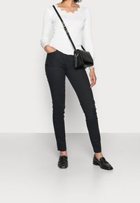 I pantaloni neri attillati presentano un design aderente e una trama liscia, abbinati a una maglietta bianca a maniche lunghe con bordi scallop e una borsa a tracolla.