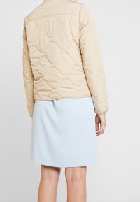 Beige prošívaná bunda s vlnitým vzorem, dlouhé rukávy; spárována se světle modrou sukní, pozorováno zezadu. Hladká textura.