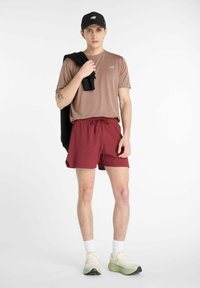 Brown athletic shirt with logo, red shorts, and light green running shoes. Black cap and sweater draped over shoulder. White background.