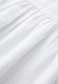 Biely textil s jemnou štruktúrou a nariasenými detailmi. Materiál pôsobí ľahko a jemne zavesený, ukazujúc hladký povrch.