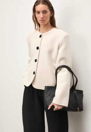 Veste structurée couleur crème avec boutons noirs, associée à un sac à main noir tissé avec poignées en cuir et fermeture éclair.