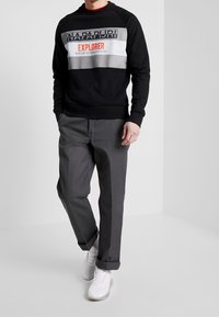 Sudadera negra con el gráfico "NAPAPIJRI EXPLORER" y diseño a rayas, combinada con pantalones grises y zapatillas blancas, mostrando un estilo casual.
