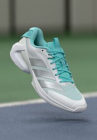 Tirkizne sportske cipele s mrežastim gornjim dijelom, srebrnim detaljima i zakrivljenim dizajnom potplata. Imaju vezice i istaknuti logo u obliku tri pruge.