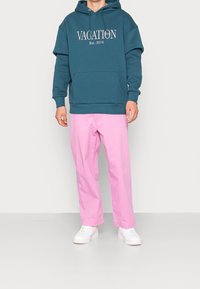 Felpa oversized color te verde acqua con la scritta "VACATION" e tasca a marsupio, abbinata a pantaloni dritti rosa chiaro e sneakers bianche.
