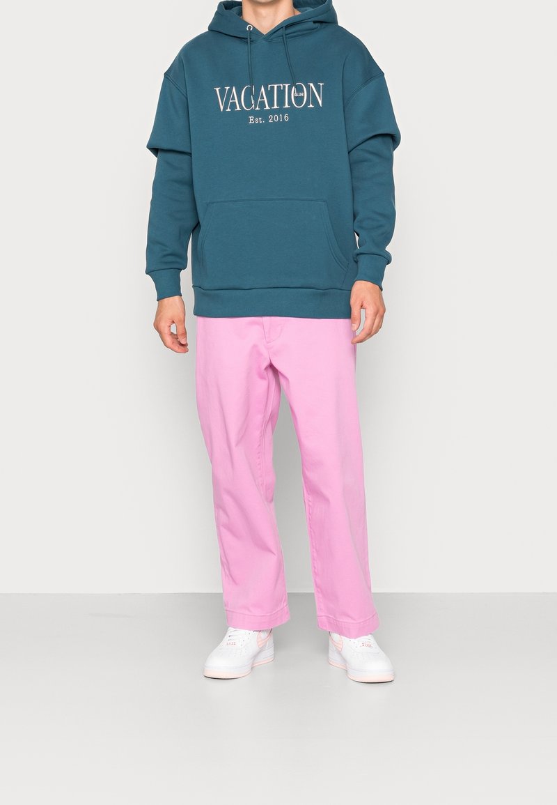 Felpa oversized color te verde acqua con la scritta "VACATION" e tasca a marsupio, abbinata a pantaloni dritti rosa chiaro e sneakers bianche.