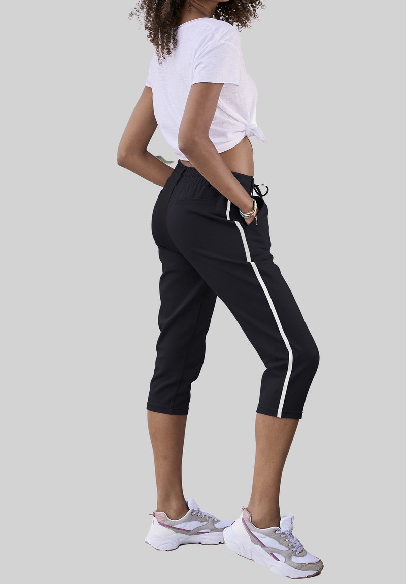 Pantalon de sport court noir avec bandes blanches sur les côtés, taille élastique et cordon de serrage. Associé à des baskets sportives blanches et grises.