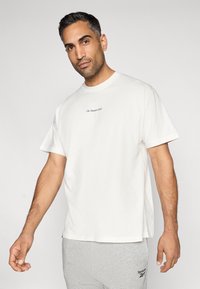 T-shirt branco em algodão com gola redonda e mangas curtas, apresentando um pequeno logotipo em azul-marinho no peito. Textura suave e ajuste descontraído.
