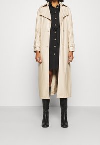 Beige long trench coat made of smooth leather, featuring a large collar, double-breasted closure, and buttoned cuffs. Worn over a black dress.