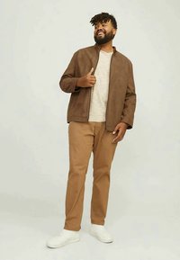 Man wearing brown jacket over beige shirt, tan pants, and white sneakers, standing and smiling against a plain light background.