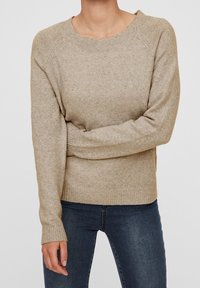 Beiger Strickpullover mit Raglanärmeln, rundem Ausschnitt und gerippten Bündchen. Weiche Textur mit subtilm Muster, kombiniert mit dunklen Jeans.