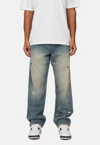 Lichtblauwe, versleten denim jeans met grote scheuren en een relaxte pasvorm. Draagt een wit T-shirt en witte sneakers. Subtiele vervaging en textuur.