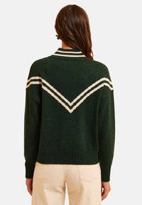 Maglione verde lavorato a maglia con un motivo a zig-zag bianco sulla schiena, caratterizzato da un colletto e polsini a coste. Il design è rilassato e leggermente oversize.
