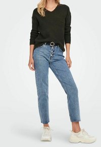 Pull en tricot noir, jeans taille haute bleu clair avec des boutons sur le devant, et baskets blanches avec des accents crème. Design simple sans motifs.