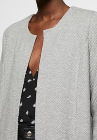 Šedý žíhaný kabát s kulatým výstřihem bez zapínání. Nositelný přes černý top s bílými puntíky.