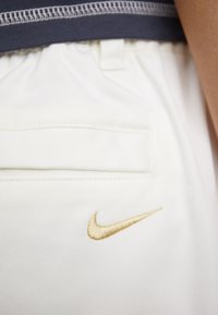 Bele športne hlače bele barve z zlatim vezenim logotipom Nike na spodnjem levem robu. Pas je izdelan iz temno modrega blaga. Gladka tekstura.