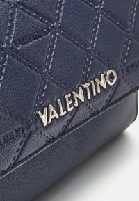 Detailní záběr na tmavě modrou prošívanou kabelku se stříbrným logem "VALENTINO" a texturovanými koženými detaily.