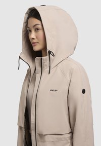 Beige waterdichte jas met een grote capuchon, een ritssluiting aan de voorkant en zwarte accenten. Heeft een logo op de borst en een textuur afwerking.