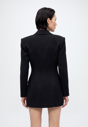 Un mannequin photographié de dos porte une robe blazer noire cintrée et tourne la tête sur le côté devant un fond blanc uni.