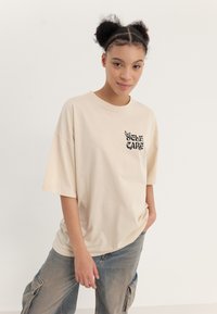 Beige oversized t-shirt made of cotton, featuring a black "SELF CARE" print and butterfly graphic on the left chest area.
