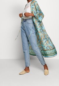 Bloemige kimono in blauw en goud, met een losse pasvorm, open voorkant en lange mouwen. Gecombineerd met lichtblauwe spijkerbroek en beige instapschoenen.