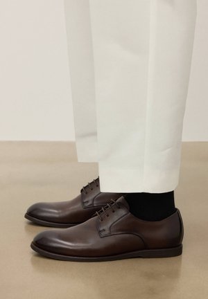 Brūnas ādas kleitu kurpes, valkātas ar melnām zeķēm un baltām biksēm, stāvot uz gluda bēša grīdas.