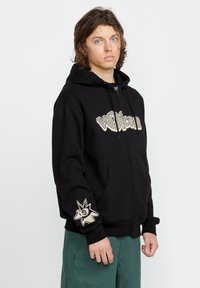 Haut de sweat à capuche zippé noir avec un grand logo beige sur le devant et un graphique d'étoile dorée sur la manche gauche. Comprend une capuche avec cordon de serrage et des poches avant.