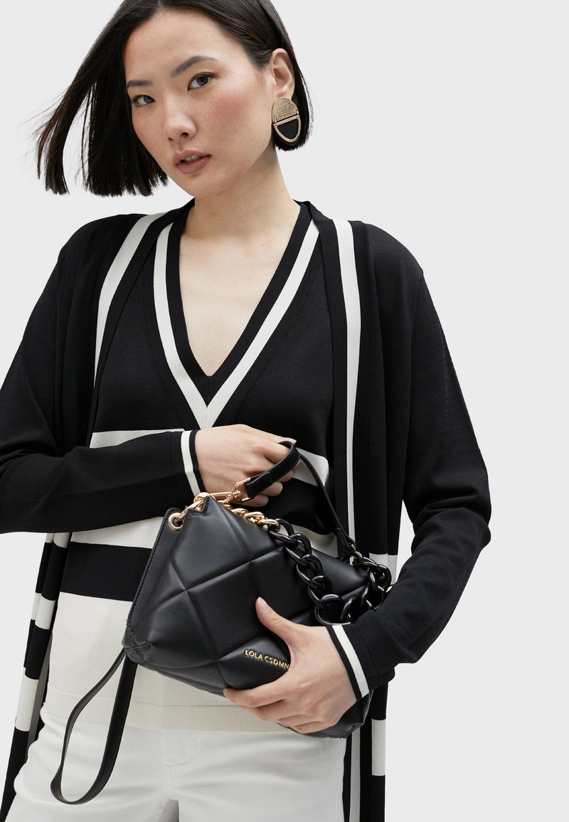 Black quilted handbag with gold accents and chain handle, held by a person in a black and white striped cardigan.