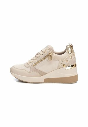 Zapatilla deportiva beige con un acabado texturizado, que presenta una cremallera lateral, acentos dorados y un tacón en cuña. Diseñada con tachuelas decorativas en la parte posterior.