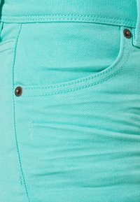 Denim kangas värvitud akvamarinitoonis, millel on sile tekstuur, nähtavad õmblused ja väike esikott, millel on pronksist nupuga detail.