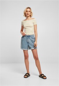 Creme, off-shoulder åtsittande topp kombinerad med ljusblå, uppvikta denimshorts. Modellen bär svarta sandaler. Enkel, avslappnad kläddesign.