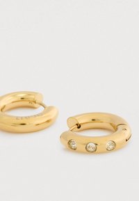 Χρυσά κρίκοι σκουλαρίκια, ένα εκ των οποίων έχει τρία ενσωματωμένα διαυγή πετράδια, τοποθετημένα σε μια ανοιχτή επιφάνεια.